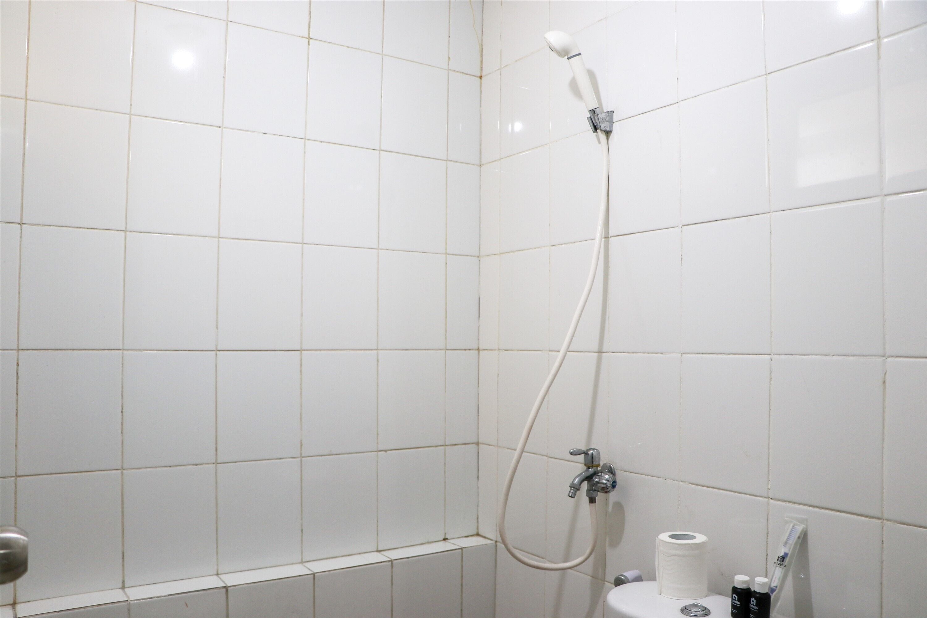 Um banheiro com paredes e piso cobertos por azulejos brancos, chuveiro com ducho manual, vaso sanitário e pia.