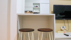 A kitchen area with a TV mounted on the wall, a countertop with a kettle, and two stools. - Best Deal And Well Furnished 2Br Bassura City Apartment (Jakarta)