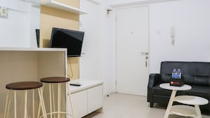 A room with a white desk, two wooden stools, a black leather sofa, a small white table with a bottle of water, and a television mounted on the wall. - Best Deal And Well Furnished 2Br Bassura City Apartment (Jakarta)