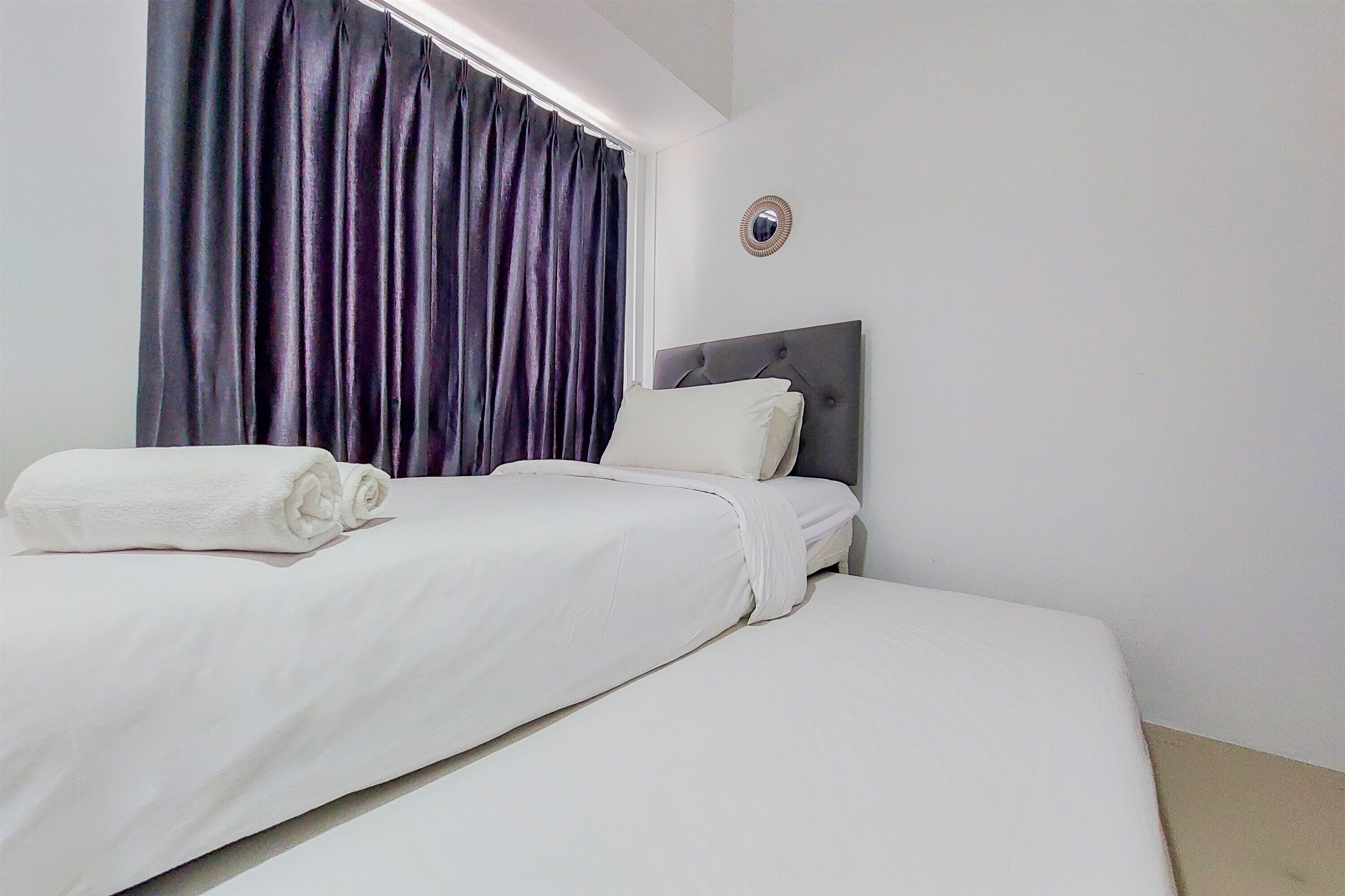 A neatly made bed with white linens and a rolled towel, a dark curtain partially covering a window, and a round mirror on the wall.