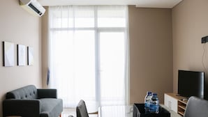 A modern living room with a sofa, a small round table, a flat-screen TV, and a large window with sheer curtains. - Modern And Compact Designed 1Br At Ciputra International Apartment (Jakarta)