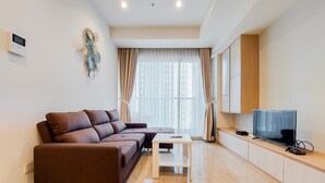 Una sala de estar moderna con un sofá marrón, una mesita blanca pequeña, un televisor sobre un mueble de madera y una ventana con cortinas translúcidas.