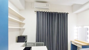 A modern, well-lit room with a white desk, a grey chair, a grey curtain, and a small black box labeled 'travelio'. - Cozy Stay Studio At Mataram City Apartment (Ngaglik)