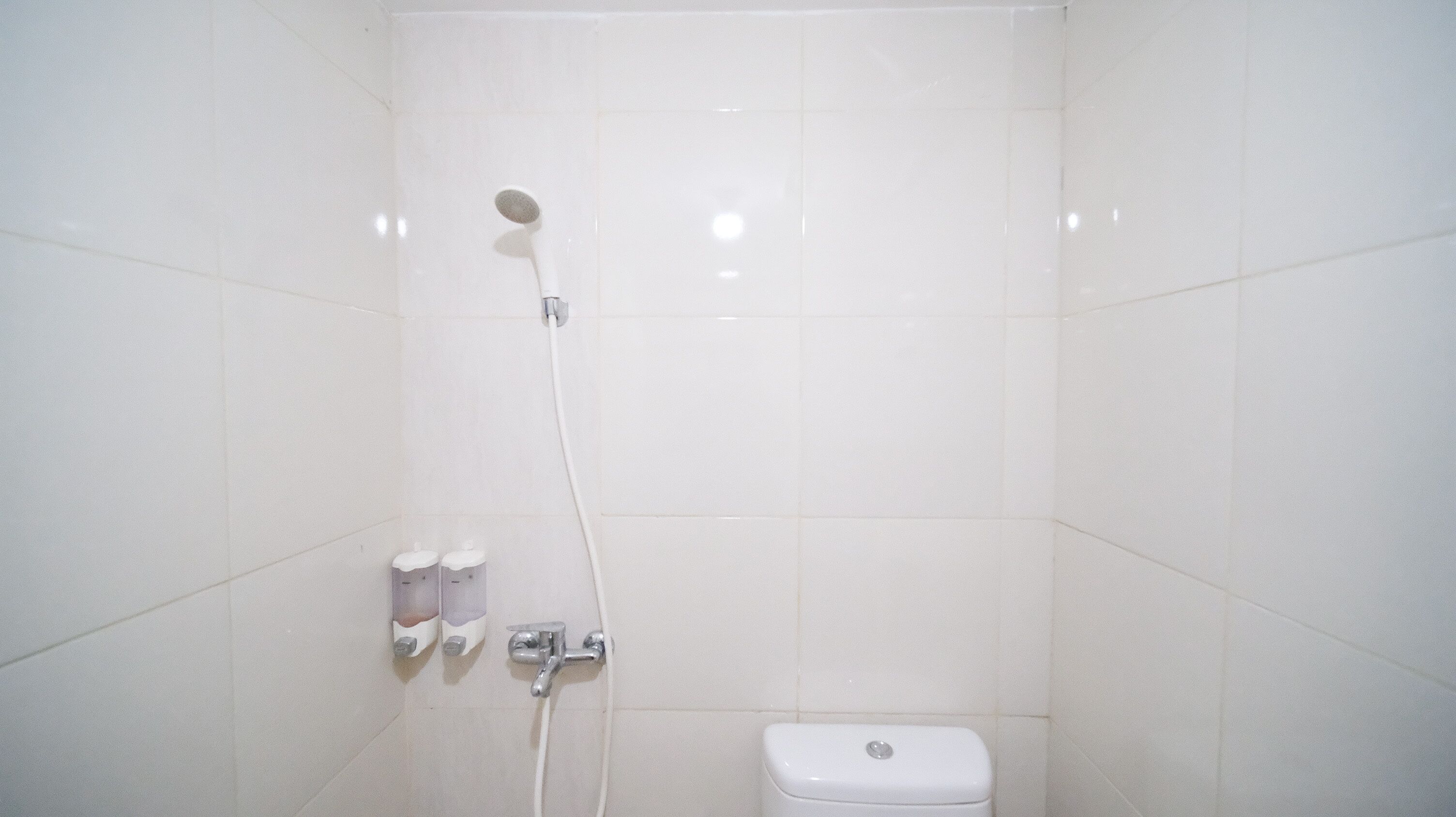 A small, white-tiled bathroom with a shower, toilet, and two soap dispensers.