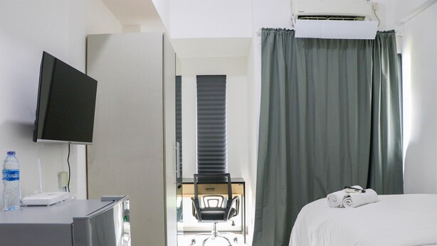 A hotel room with a bed, a desk with a TV, a chair, and a window with curtains. - Cozy And Comfort Stay Studio Sayana Bekasi Apartment (Tarumajaya)