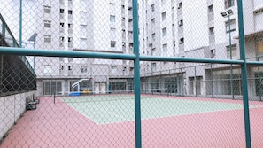 Ein Tennisplatz im Freien, umzäunt mit einem Maschendrahtzaun, mit einem mehrstöckigen Gebäude im Hintergrund.