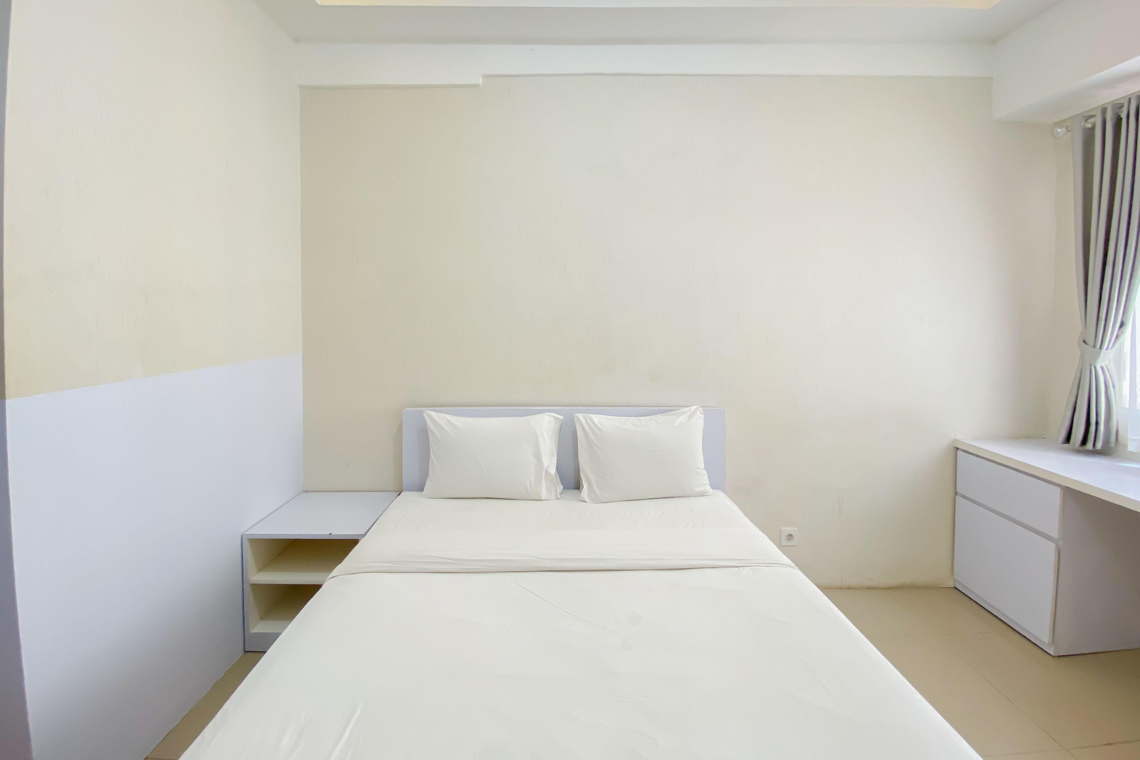 A minimalist hotel room with a single bed, a small bedside table, a nightstand, and a desk with a chair.