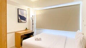 A modern hotel room with a bed, a desk, a chair, a lamp, and a painting on the wall. - Spacious And Homey 2Br At Meikarta Apartment (Cikarang)
