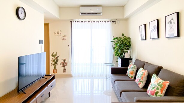 A modern living room with a flat-screen TV, a sofa with colorful cushions, a coffee table, and a large potted plant. - Spacious And Homey 2Br At Meikarta Apartment (Cikarang)