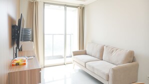 A modern living room with a sofa, a flat-screen TV mounted on the wall, a wooden desk with a computer and a bag of chips, and a sliding glass door leading to a balcony. - Wonderful And Homey 2Br At Gold Coast Apartment (Jakarta)