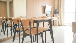 A modern hotel room with a dining area featuring a wooden table and chairs, a flat-screen TV mounted on the wall, and a large window with curtains. - Wonderful And Homey 2Br At Gold Coast Apartment (Jakarta)