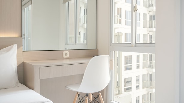 Ein modernes Hotelzimmer mit einem Bett, einem weißen Stuhl, einem großen Spiegel und einem Fenster mit Blick auf ein Gebäude.