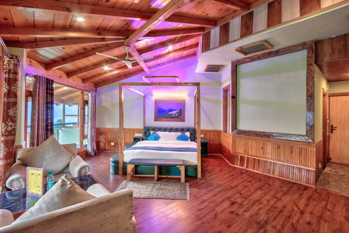 A spacious room with a large bed, wooden ceiling, and a view of the mountains through a window.