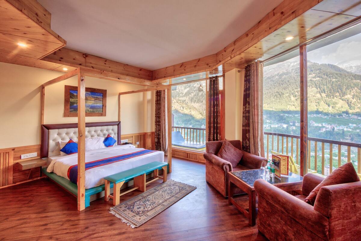A bedroom with a large bed, a wooden bench, a sofa, and a view of a mountainous landscape.