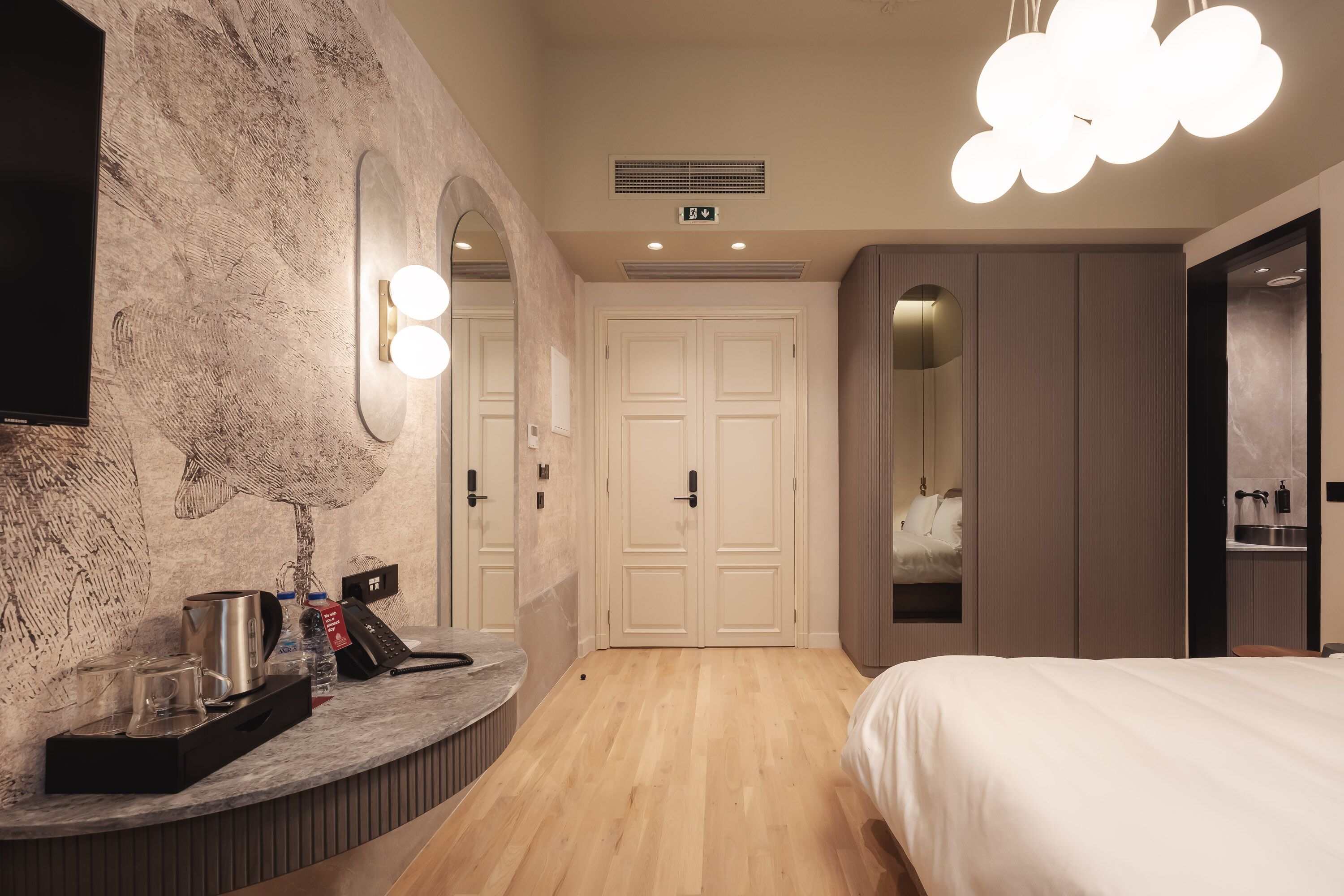 Una habitación de hotel moderna con piso de madera, una cama, un tocador con espejo e iluminación, una estación de café y un armario.
