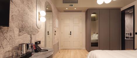 Una habitación de hotel moderna con piso de madera, una cama, un tocador con espejo e iluminación, una estación de café y un armario.