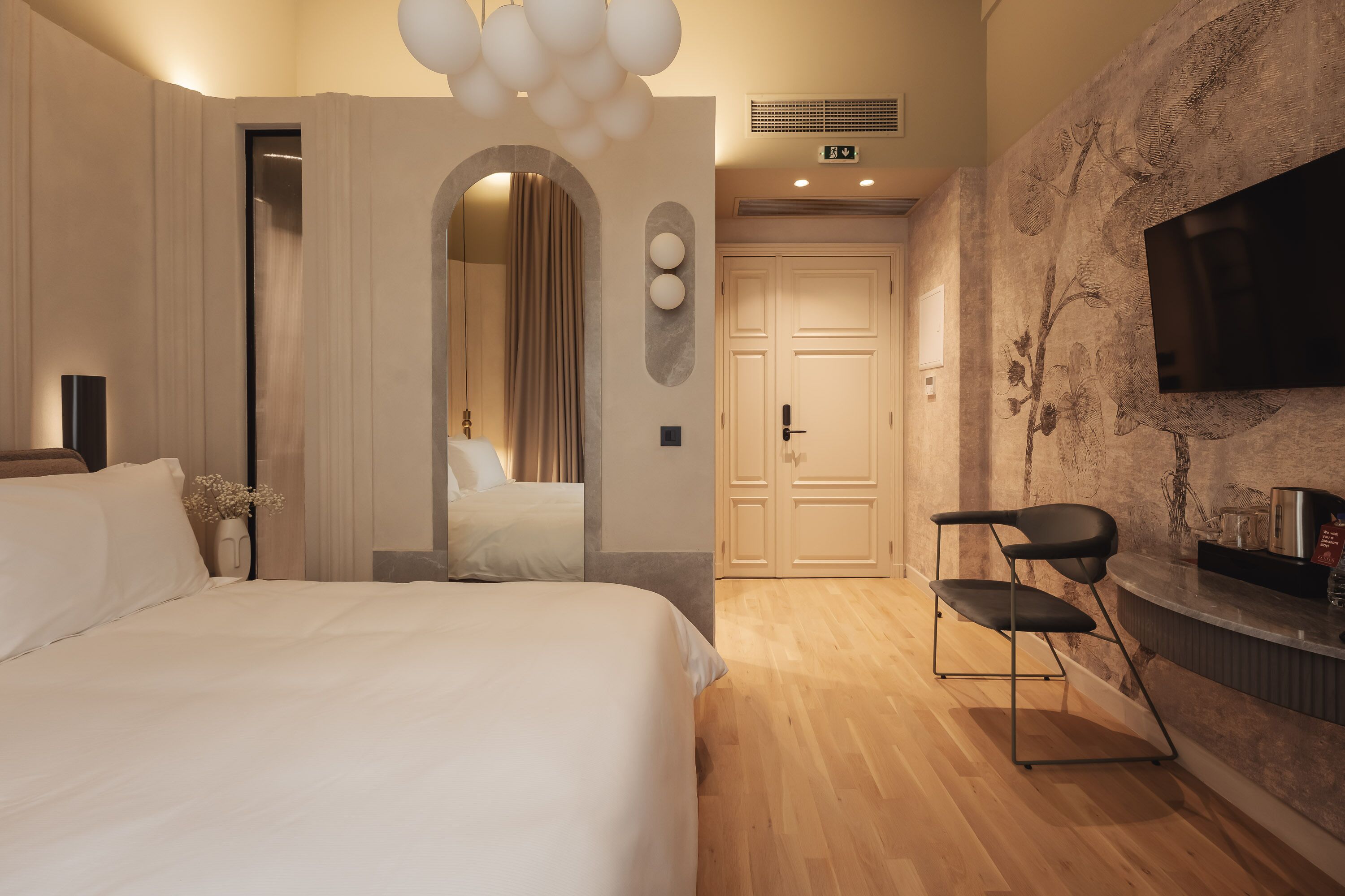 Habitación de hotel con una cama, una silla, un televisor montado en la pared y una puerta que da a otra habitación.