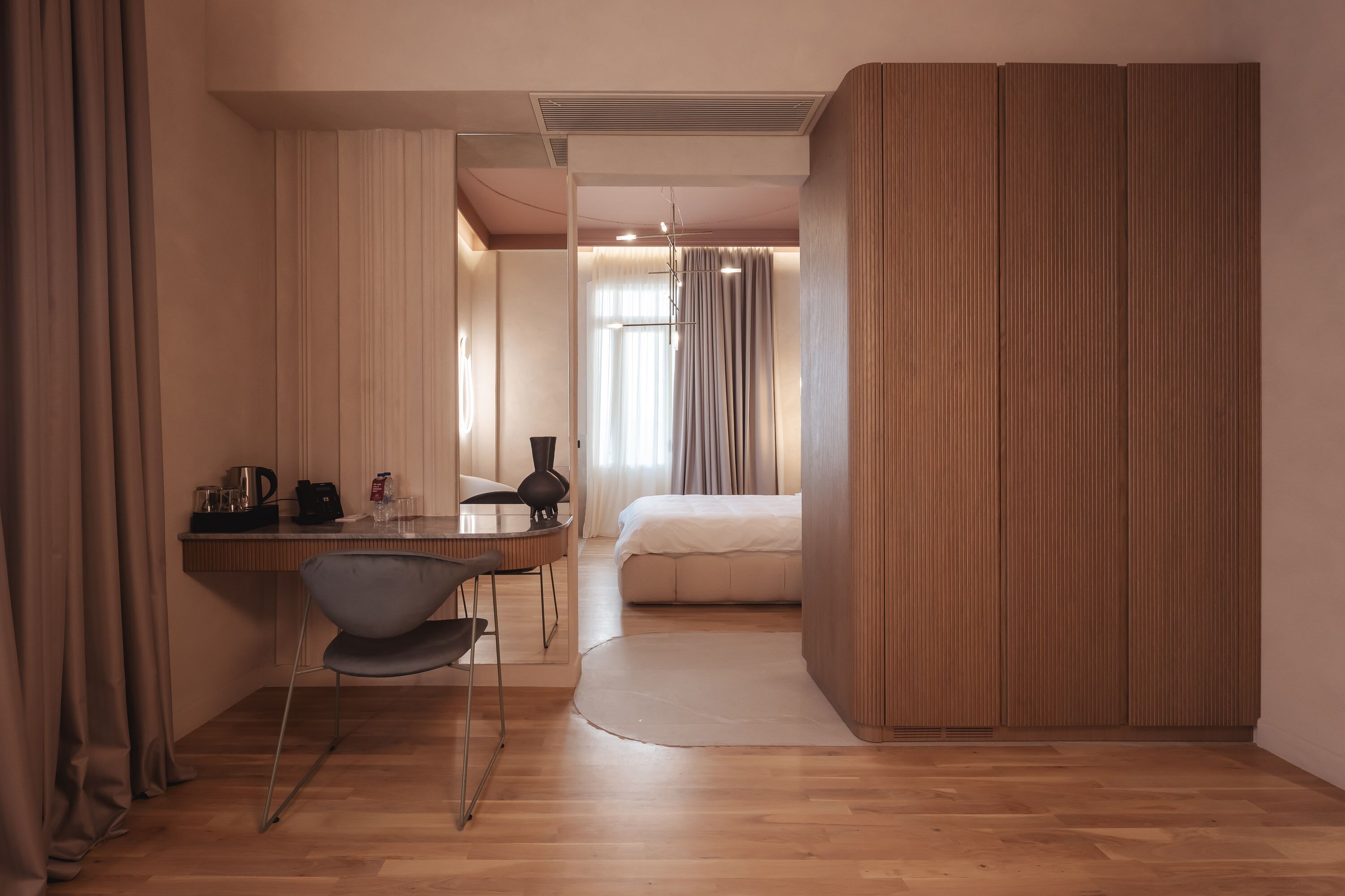 A modern hotel room with a wooden floor, a bed, a desk with a chair, and a large window with curtains.