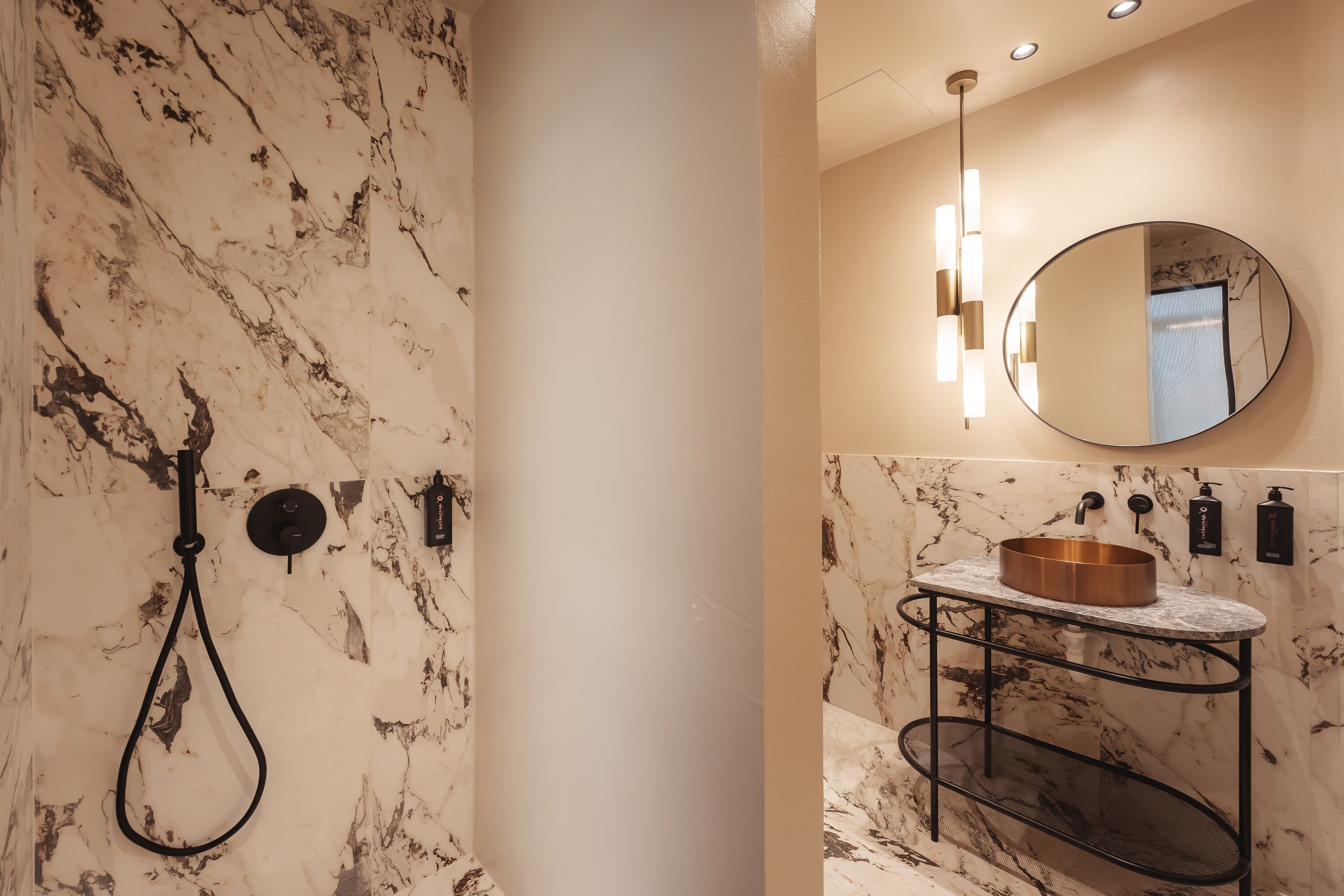 A bathroom with a marble wall, a round sink with a copper basin, and a black metal shelf.