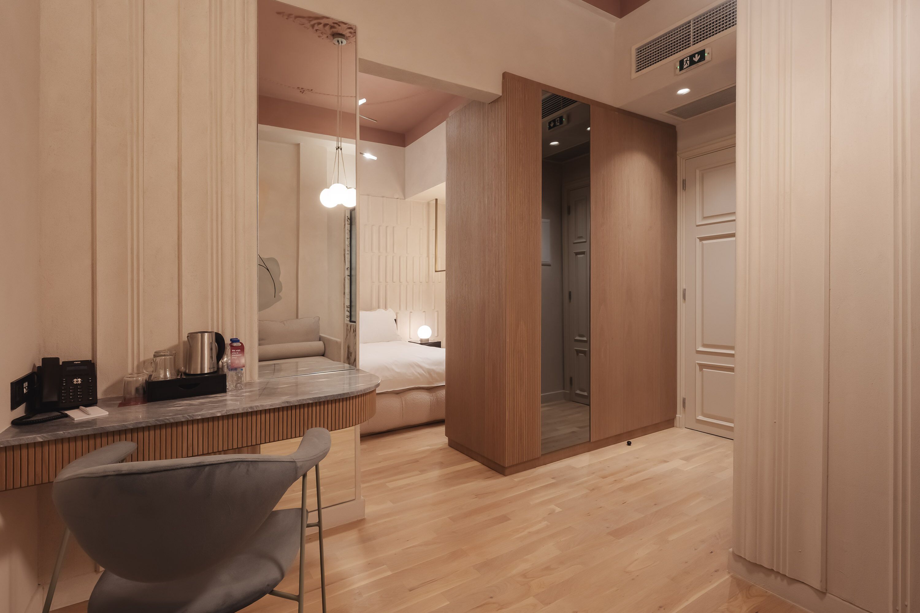 Une chambre d’hôtel moderne avec un plancher en bois, un lit doté d’une tête de lit blanche, un petit coin cuisine équipé d’une machine à café et un grand miroir.