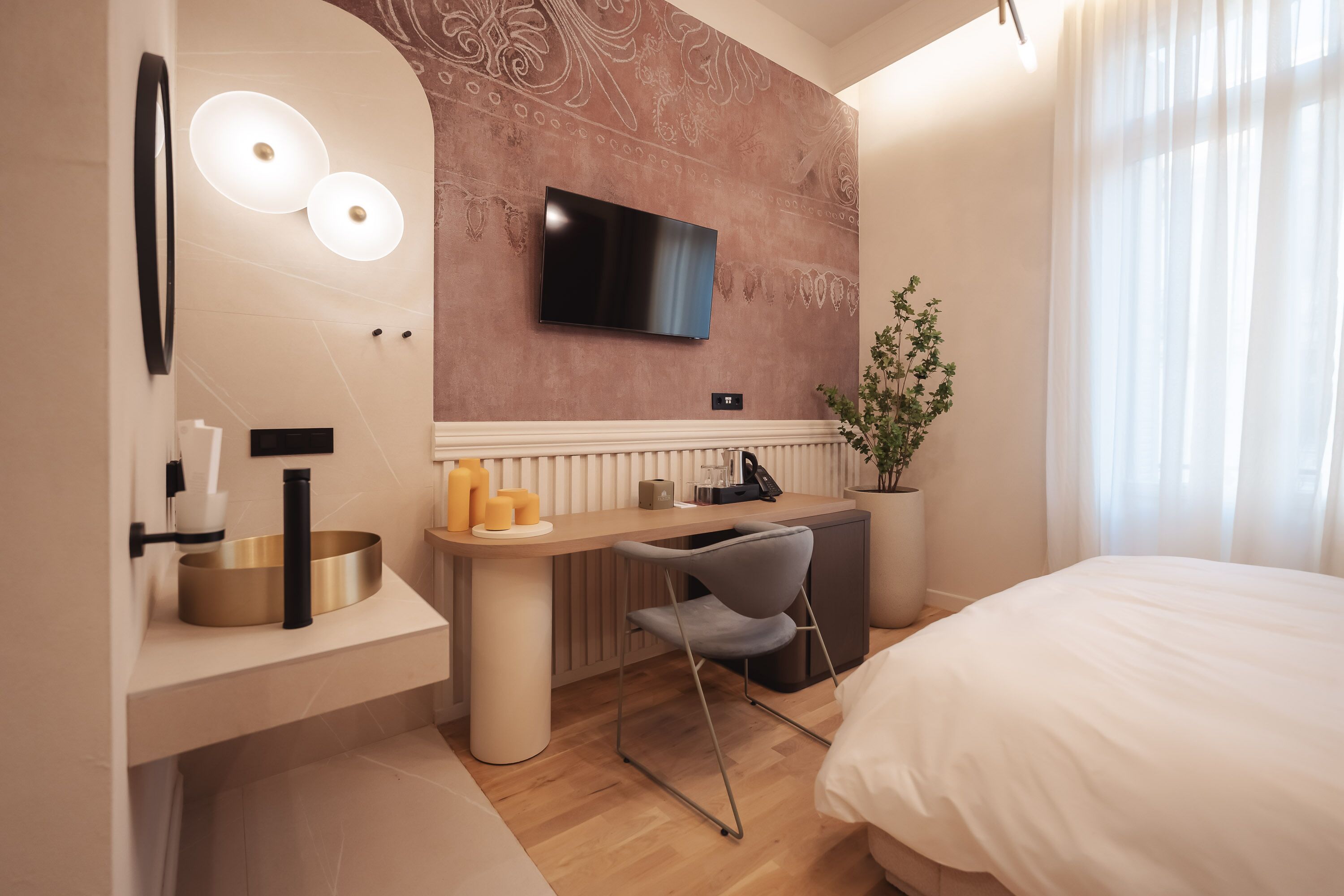 Una habitación de hotel moderna con una cama, un escritorio con una silla, un televisor montado en la pared, una planta grande en maceta y un baño con lavabo y espejo.