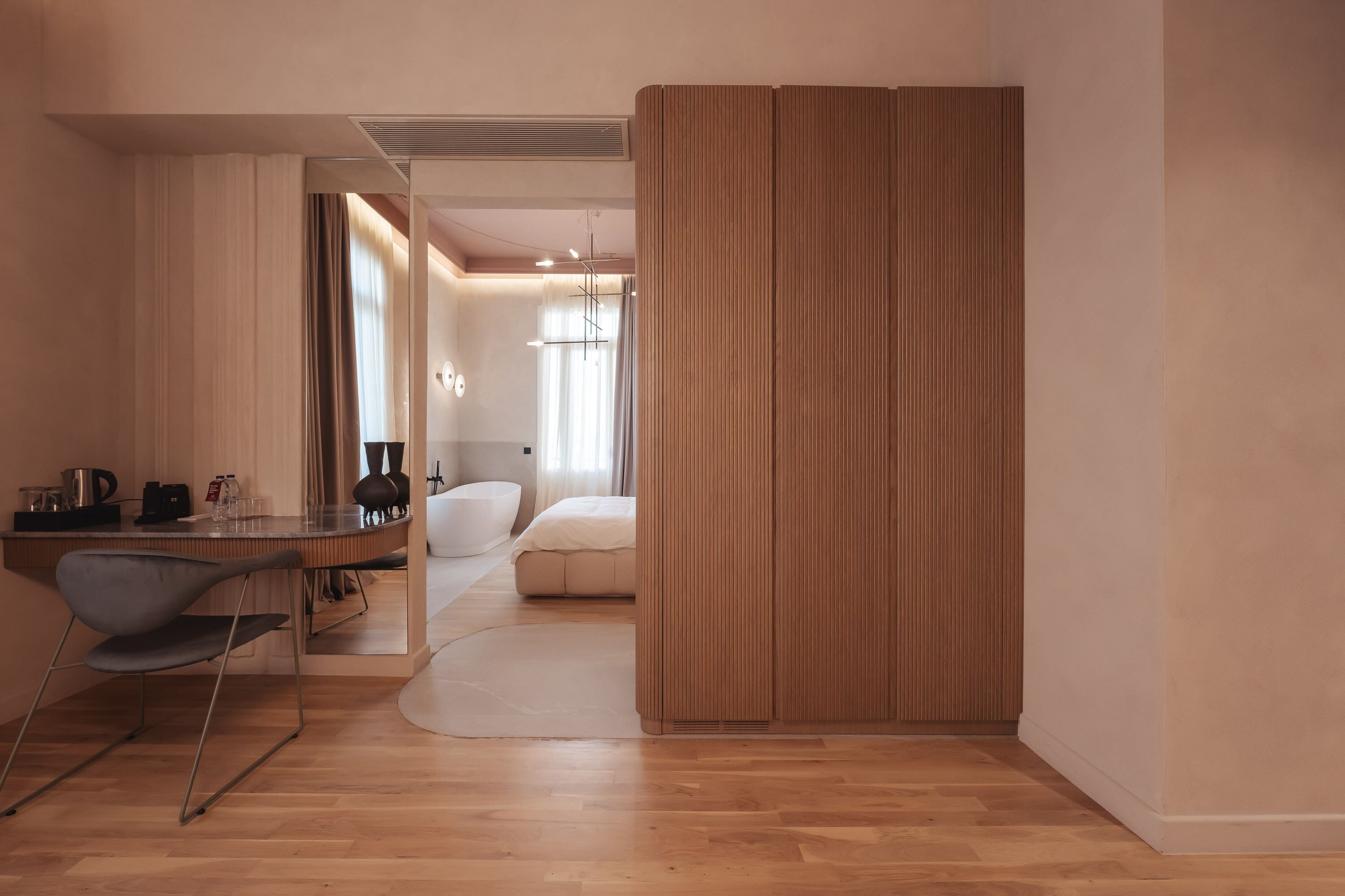 A modern hotel room with a wooden floor, a desk with a chair, a bed, and a sliding door.