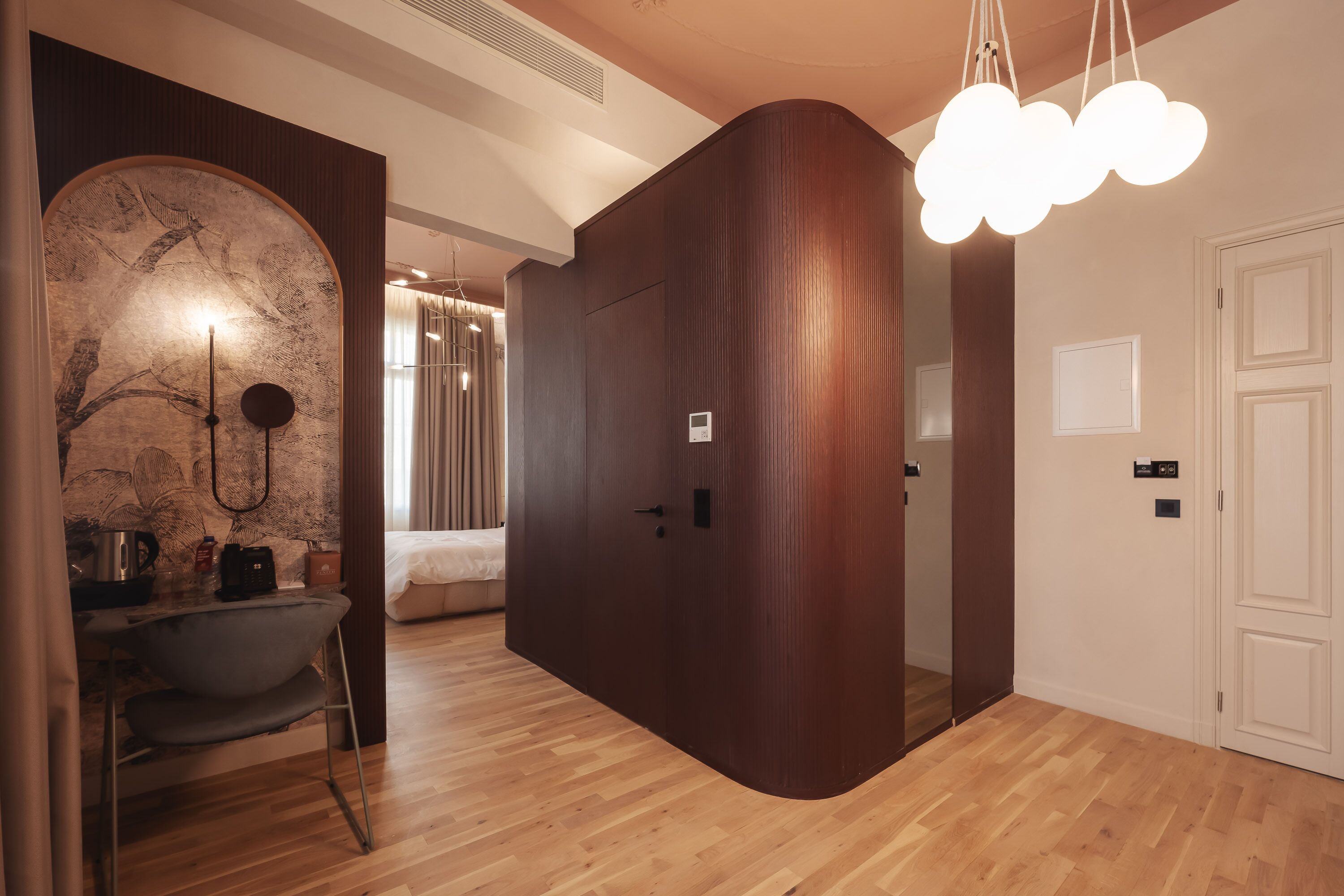 A modern hotel room with a wooden floor, a curved wall with a decorative mural, a dining area with a chair and table, and a unique cloud-shaped light fixture.