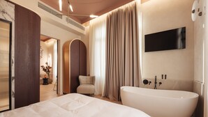 A modern hotel bathroom with a large bathtub, a flat-screen TV, and a seating area with a chair and a small table.