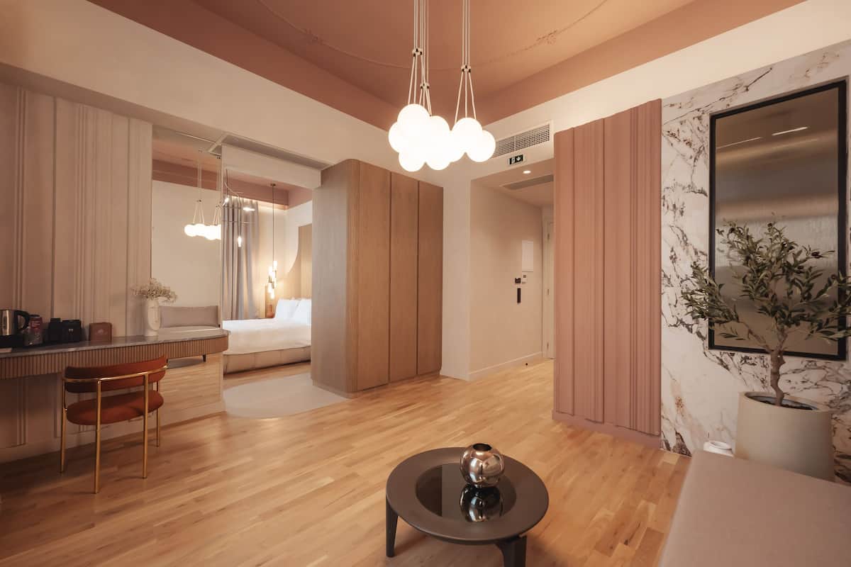 Una habitación de hotel moderna con suelo de madera, una mesa redonda con un florero decorativo, un sofá, un comedor con una silla y un espejo grande.