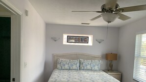 A bedroom with a bed, a ceiling fan, a nightstand with a lamp, and a window with blinds.