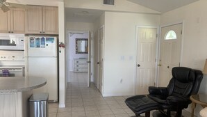 A modern kitchen with a refrigerator, microwave, and a small dining area. There is a black leather chair and a trash can in the living area.