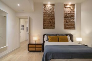 A bedroom with a bed, two bedside lamps, a nightstand, a wall-mounted artwork, and a door leading to another room.