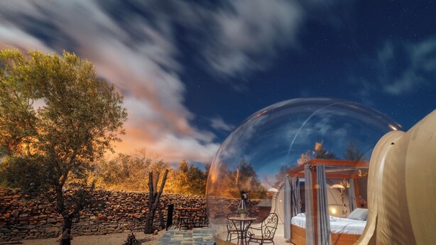 A transparent dome with a bed and seating area inside, surrounded by a stone wall and trees. - Hotel Zielo de Levante (Tirig)