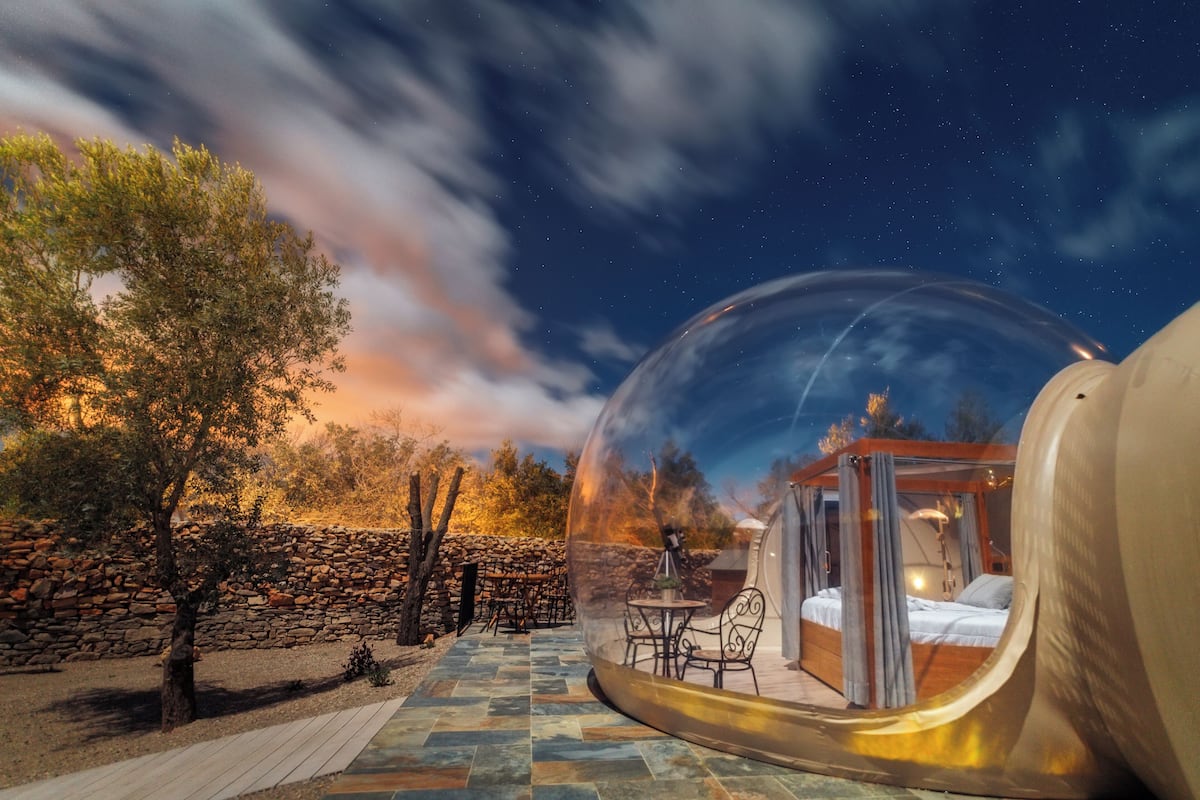 A transparent dome with a bed and seating area inside, surrounded by a stone wall and trees.