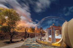 A transparent dome with a bed and seating area inside, surrounded by a stone wall and trees. - Hotel Zielo de Levante (Tirig)