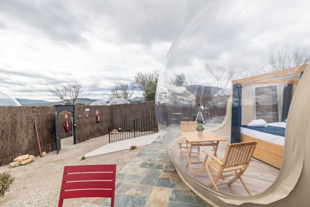 A transparent dome room with a bed, wooden table, and chairs.