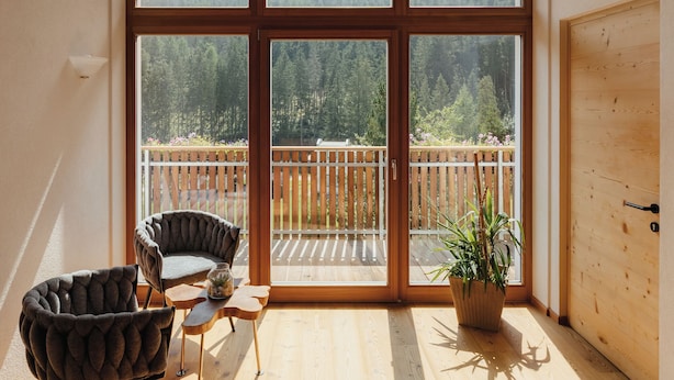 Una habitación con ventanas amplias, techo de madera, balcón, una silla, una mesita y una planta en maceta.