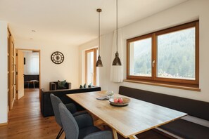 Un comedor moderno con una mesa de madera, asientos tipo banco, lámparas colgantes y vistas a un bosque a través de amplios ventanales.