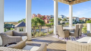 A covered outdoor area with wicker furniture, a table, and chairs. There are white pillars and a view of houses in the background. - Luxury Coastal Retreat | Heated Private Pool | Resort Style Amenities (Gulf Shores)
