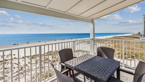 Ein überdachter Balkon mit Tisch und Stühlen, mit Blick auf einen Strand mit Menschen und einem klaren Himmel.