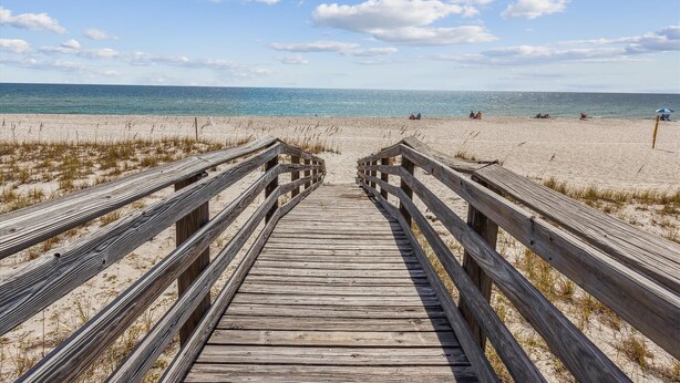 Ein hölzerner Steg führt zu einem Strand bei blauem Himmel und wenigen Personen in der Ferne.