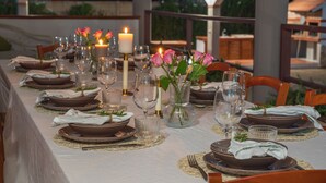 Una larga mesa de comedor preparada para una comida, con velas, platos, copas y un florero con flores.