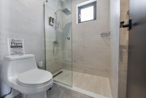 A modern bathroom with a glass-enclosed shower, a toilet, and a sign advising against flushing certain items down the pipes.