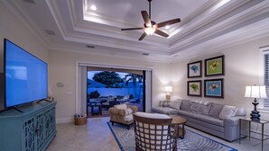 A spacious living room with a flat-screen TV, a ceiling fan, a sofa, a coffee table, and a wicker chair.