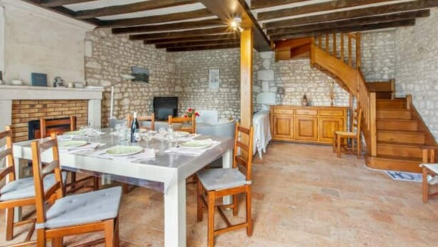 A spacious dining area with a long table set for a meal, surrounded by wooden chairs. A stone fireplace and a kitchen area with wooden cabinets and a staircase are visible in the background. - Vacances Gites Assay Maisons Chaleureuse (Assay)