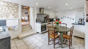 A modern kitchen with a stone wall, a dining area with a round table and chairs, and a visible staircase leading to another level. - Vacances Gites Assay Maisons Chaleureuse (Assay)