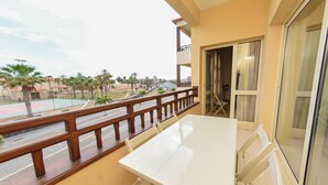 Un balcon avec une table et des chaises blanches, une balustrade en bois et une vue sur une route et des palmiers.