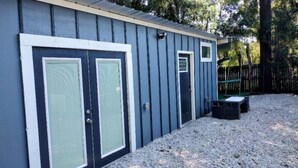 A building with two large doors and a smaller door, surrounded by gravel and trees. - Backyard Guest House Suite (Gainesville)