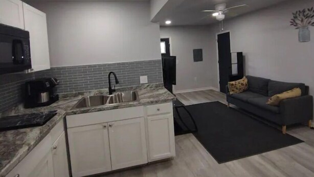 A modern kitchen with white cabinets, a granite countertop, and a black faucet. There is a gray couch with a black rug in the living area. - Backyard Guest House Suite (Gainesville)