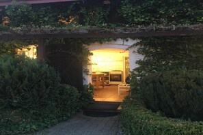 Un espacio exterior acogedor con una pérgola de madera cubierta de follaje, que da paso a un ambiente interior cálidamente iluminado con una mesa y sillas para comer.